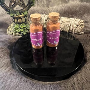 May include: Two small glass jars with cork lids filled with a red powder. The jars have purple labels with white text that reads "Nightshade & Sage" and "Red Brick Dust".