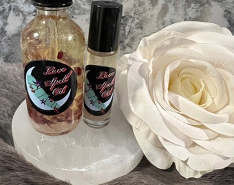 Puede incluir: Dos botellas de vidrio de Love Spell Oil con tapas negras y etiquetas con un diseño de media luna. Las botellas están sobre una piedra blanca en forma de corazón, junto a una gran rosa blanca. El aceite contiene elementos botánicos visibles.