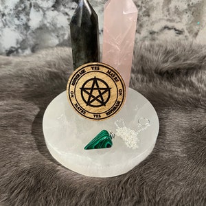 Könnte beinhalten: Ein Holz-Pendelbrett mit den Worten "Ja", "Vielleicht", "Nein" und "Neu formulieren" um einen fünfzackigen Stern. Ein grünes Malachit-Pendel hängt an einer silbernen Kette über dem Brett. Zwei Kristallspitzen, eine graue und eine rosa, stehen hinter dem Brett.