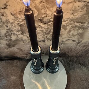 May include: Two black candles with bright flames are set in ornate holders, each adorned with a key charm. The holders sit on a round, pale stone base, creating a mystical and decorative display.