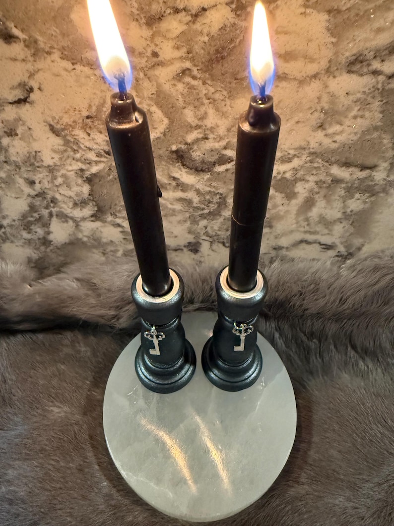 May include: Two black candles with bright flames are set in ornate holders, each adorned with a key charm. The holders sit on a round, pale stone base, creating a mystical and decorative display.