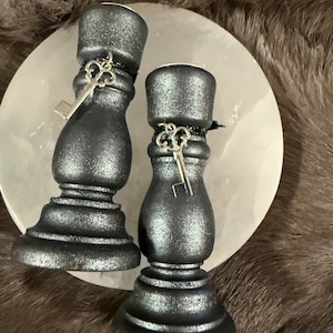 May include: Two dark gray candle holders with a metallic finish, each adorned with a small silver key charm. The candle holders are placed on a white, round surface, set against a backdrop of faux fur.