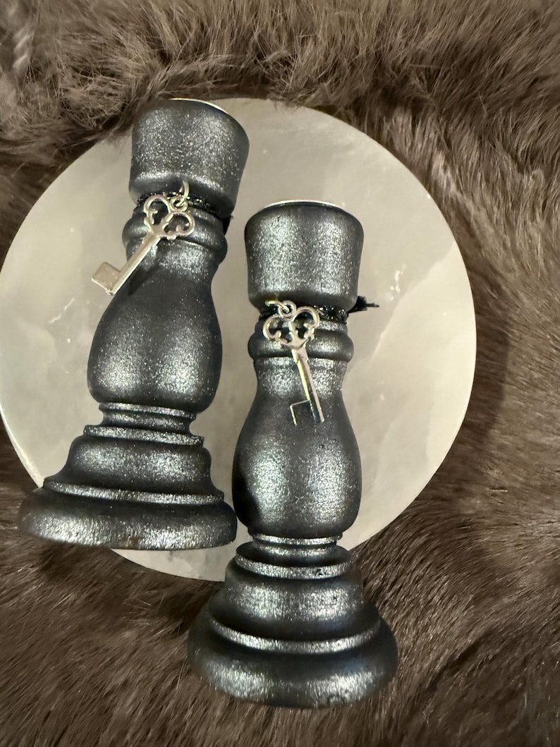 May include: Two dark gray candle holders with a metallic finish, each adorned with a small silver key charm. The candle holders are placed on a white, round surface, set against a backdrop of faux fur.