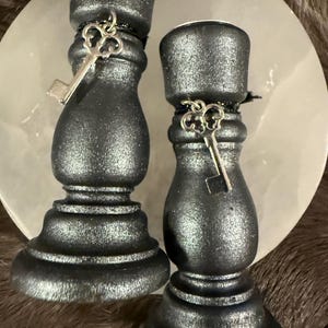 May include: Two black candle holders with a metallic sheen. Each holder has a decorative silver key charm. The candle holders are on a light-colored, round surface, with a dark, textured background.