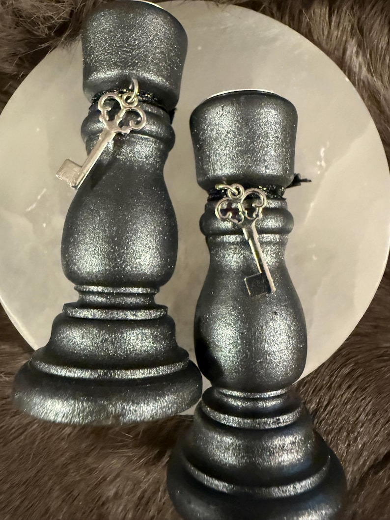 May include: Two black candle holders with a metallic sheen. Each holder has a decorative silver key charm. The candle holders are on a light-colored, round surface, with a dark, textured background.