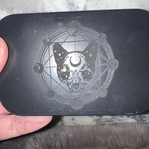 May include: Black rectangular box with a silver skull-like cat design inside a circle with a crescent moon and stars. The design is engraved into the box.