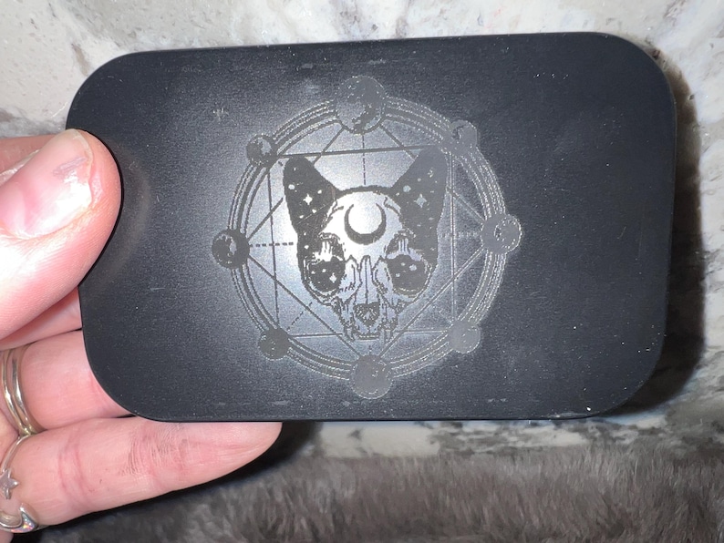 May include: Black rectangular box with a silver skull-like cat design inside a circle with a crescent moon and stars. The design is engraved into the box.