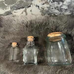 Puede incluir: Tres botellas de vidrio con tapas de corcho. Las botellas son transparentes y tienen diferentes formas y tama&ntilde;os. Las botellas est&aacute;n sobre una superficie gris y peluda.