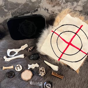 May include: A collection of various objects, including bones, a seashell, a black stone, a wooden disc, a crystal, a metal button, a metal key, a honeycomb, a metal tin with a black and white symbol, and a white piece of fabric with a red cross and black circles.