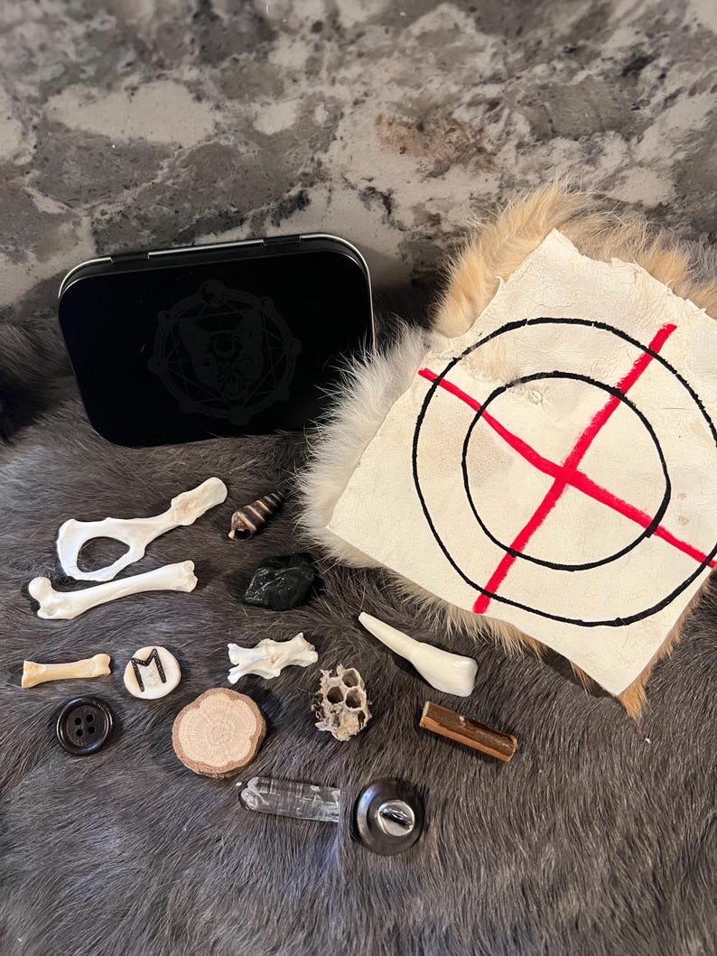 May include: A collection of various objects, including bones, a seashell, a black stone, a wooden disc, a crystal, a metal button, a metal key, a honeycomb, a metal tin with a black and white symbol, and a white piece of fabric with a red cross and black circles.