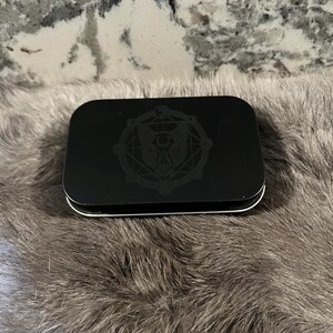 May include: A black metal tin with a circular design featuring a stylized owl with geometric patterns and the words "The Owl and the Pussycat".