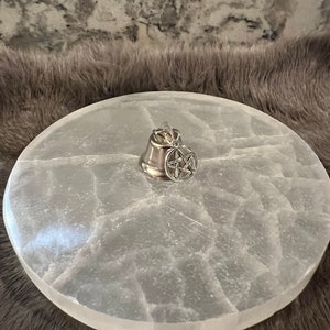 Pu&ograve; includere: Un piatto di ricarica in selenite bianco con un ciondolo a campana d'argento con un pentagramma. Il piatto ha una superficie naturale testurizzata.