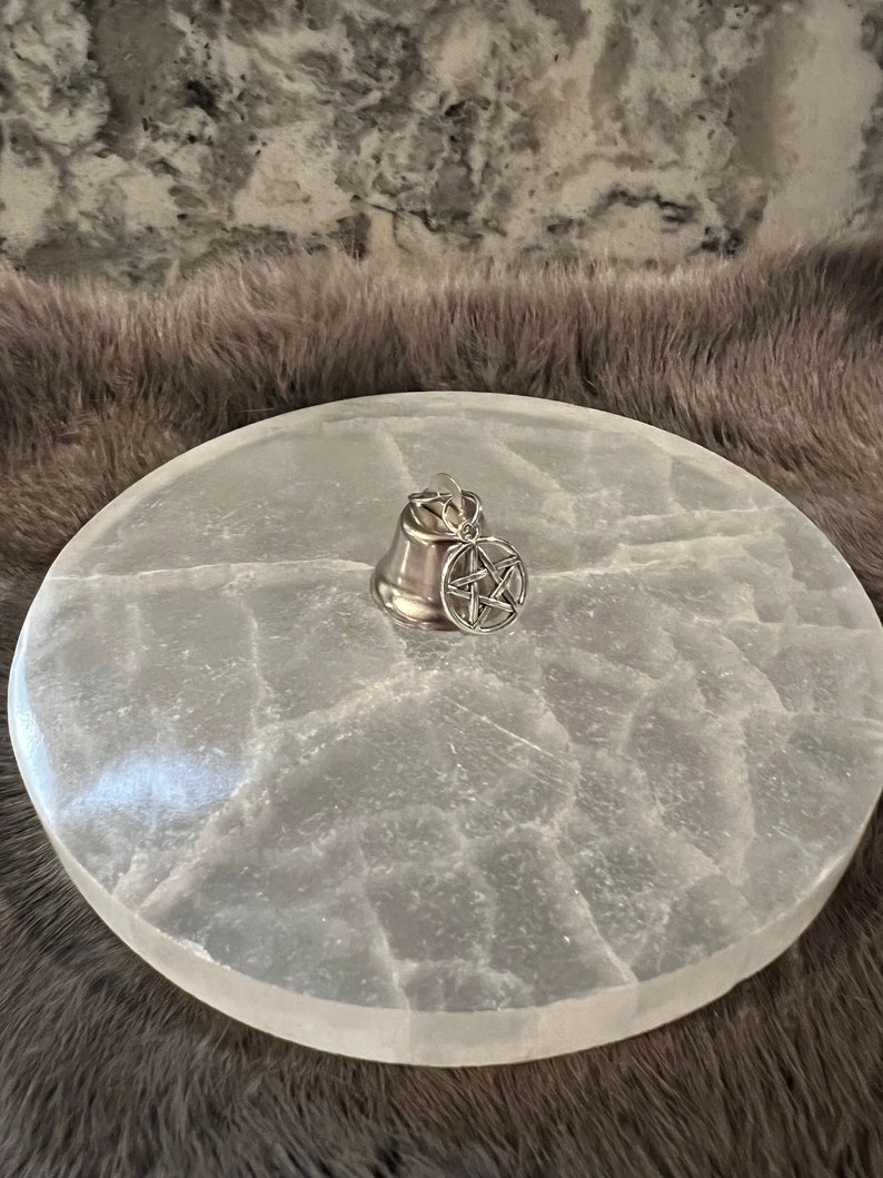 Pu&ograve; includere: Un piatto di ricarica in selenite bianco con un ciondolo a campana d'argento con un pentagramma. Il piatto ha una superficie naturale testurizzata.