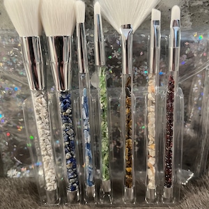 May include: Set of seven makeup brushes with clear handles filled with colorful crystals. The brushes have white bristles and are arranged in a clear plastic holder.
