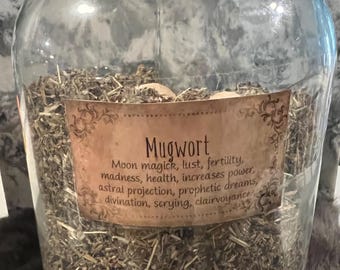 May include: A glass jar filled with dried mugwort, a type of herb used in herbalism and witchcraft. The jar has a label that reads "Mugwort" and lists its uses, including "moon magick, lust, fertility, madness, health, increases power, astral projection, prophetic dreams, divination, scrying, clairvoyance."