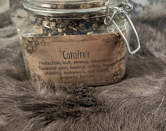 Könnte beinhalten: Ein Glasgefäß mit einem Metallverschlussdeckel, gefüllt mit getrockneten Kräutern. Das Etikett lautet "Comfrey" und listet seine Verwendung auf: Schutz, Glück, Geld, Fülle, finanzieller Gewinn, Heilung, Sicherheit, Reisen, Stabilität, Ausdauer, Immobilien, Eigentum, Vermögen.