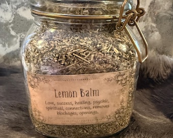 May include: A glass jar with a metal clasp lid filled with dried lemon balm. The jar has a label that reads "Lemon Balm" and lists its uses: "Love, success, healing, psychic, spiritual, connections, removes blockages, openings."