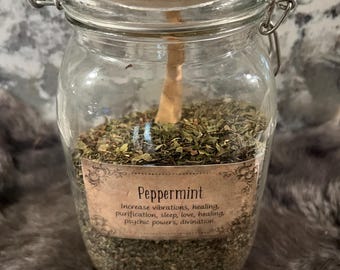 May include: A clear glass jar with a metal clasp and lid, filled with dried green herbs. A label on the jar reads "Peppermint" and lists associated properties. The jar sits on a textured, neutral-toned surface.
