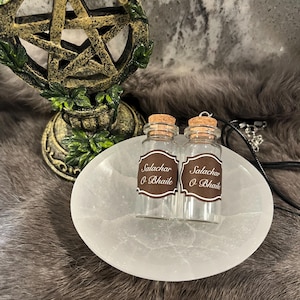 Puede incluir: Dos pequeñas botellas de vidrio con tapones de corcho, etiquetadas "Salachar O Bhaile", descansan sobre un plato ovalado blanco. Un pentagrama decorativo dorado y un cordón negro con un dije están en el fondo. El conjunto está sobre una superficie de piel sintética.