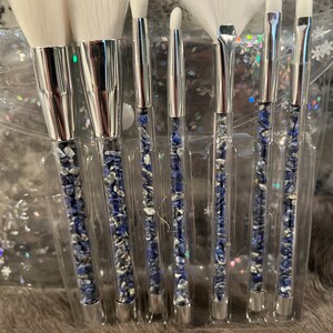 May include: Set of seven makeup brushes with clear handles filled with blue and white stones. The brushes have white bristles and silver metal ferrules.