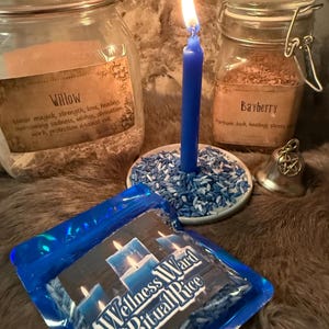 May include: A blue candle burning on a plate of blue and white rice, flanked by two glass jars labeled Willow and Bayberry. A small silver bell with a pentagram charm sits nearby, and a blue plastic pouch labeled Wellness Ward Ritual Rice.