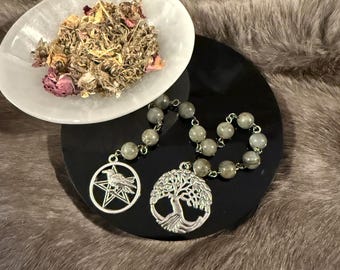 May include: A beaded necklace with two silver charms, one a tree of life and the other a bird within a star. A white bowl holds dried herbs and flowers. The items rest on a black surface and a faux fur background.
