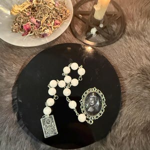 May include: A white beaded rosary with a silver pendant and a cameo brooch rests on a black circular surface. A white bowl with dried herbs and flowers sits nearby, along with a lit candle in a pentagram holder. The items suggest a spiritual or metaphysical theme.