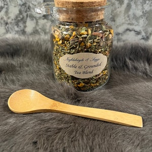May include: A glass jar with a cork lid filled with a blend of dried herbs and flowers. The jar has a label that reads "Nightshade & Sage Stable & Grounded Tea Blend". A wooden spoon is also pictured.