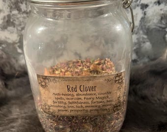 Könnte beinhalten: Ein Glasgefäß gefüllt mit getrockneten Rotkleeblüten. Das Gefäß hat einen Metallverschluss und ein Etikett mit der Aufschrift "Red Clover" und einer Auflistung seiner Verwendungsmöglichkeiten, darunter Schutz vor Hexerei, Fülle, Gegenzauber, Exorzismus, Feenmagie, Fruchtbarkeit, Treue, Glück, Hexenbrechen, Liebe, Glück, Gedächtnis, Geld, Macht, Wohlstand und Schutz.