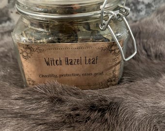 May include: A glass jar filled with dried witch hazel leaves. The jar has a metal clasp and a label that reads "Witch Hazel Leaf" and "Chastity, protection, eases grief."