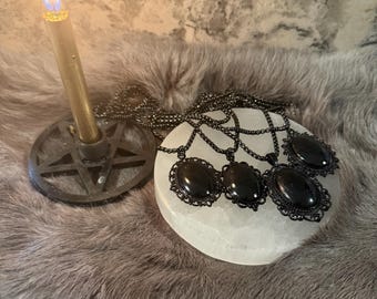 May include: A lit gold candle in a black pentagram holder next to a white stone with several black oval pendant necklaces with ornate frames. The items are on a gray faux fur surface.