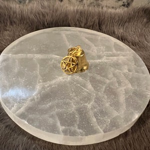Pu&ograve; includere: Una campana dorata con un ciondolo a pentagramma appeso, appoggiata su un piatto di ricarica in selenite bianca.