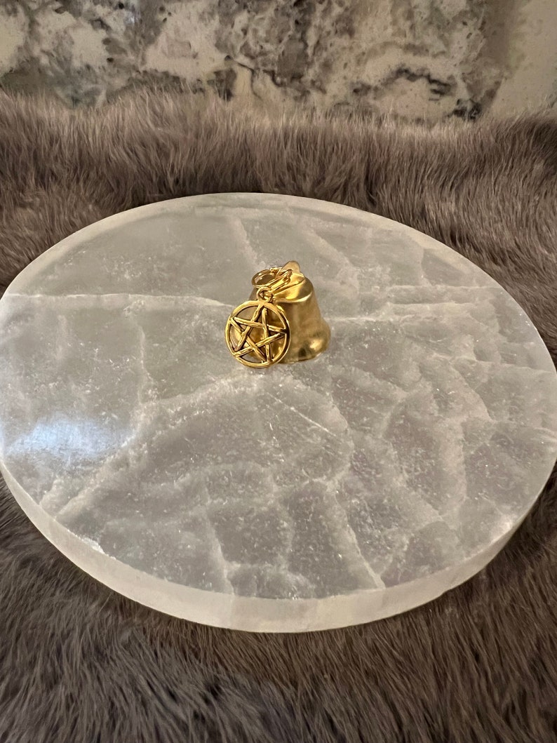 Pu&ograve; includere: Una campana dorata con un ciondolo a pentagramma appeso, appoggiata su un piatto di ricarica in selenite bianca.