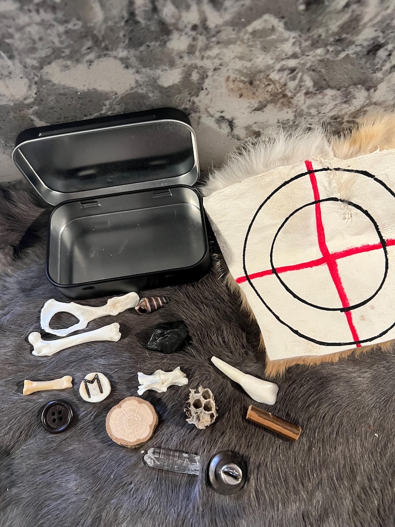 May include: A black metal tin with a variety of small objects, including bones, a shell, a crystal, a button, a wooden disc, a honeycomb, a piece of wood, a crystal, a button, and a piece of wood. The objects are arranged on a white and brown fur surface. A white target with a red bullseye is in the background.