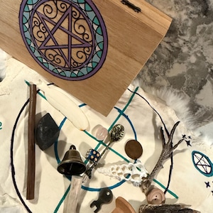 May include: A wooden box with a painted purple and teal pentacle design sits above a white cloth featuring a blue circular pattern. The cloth holds a collection of items including a brass bell, a wooden spool, a shell, a die, and a bird foot.