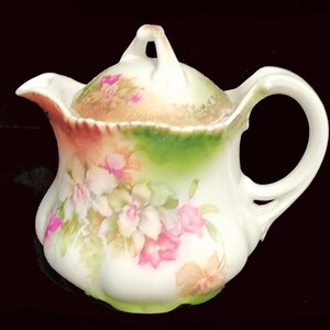 May include: A white porcelain creamer with a floral design. The creamer has a lid and a handle. The flowers are pink, white, and yellow.