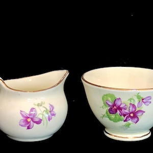 May include: Cream-colored creamer and sugar bowl set. The creamer has a handle and spout, while the bowl is round. Both pieces are decorated with purple violet floral designs and gold trim. A vintage tea set.