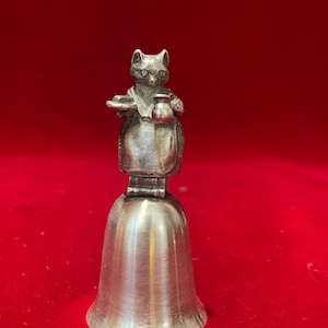 May include: A silver bell with a figurine of a cat holding a plate and a pitcher on top. The cat is wearing a dress and has a white collar.