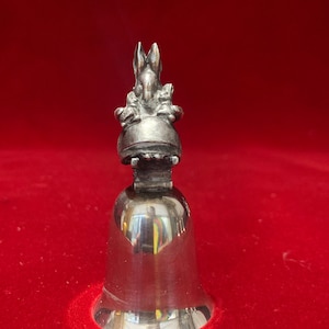 May include: A silver bell with a rabbit figurine on top. The rabbit is sitting on a round base and has its paws up. The bell is on a red surface.