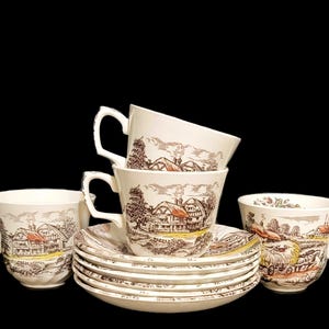 May include: Set of six vintage teacups and saucers with a brown and white cottage scene design. The cups and saucers are stacked and arranged on a black background.