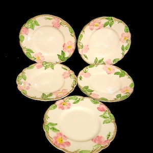May include: Five off-white ceramic plates with scalloped edges. Each plate is decorated with pink floral and green leaf designs. The plates have a gold trim around the edges. The plates are arranged on a black background.