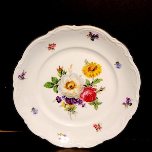 May include: White porcelain plate with a gold rim and a floral design. The flowers are yellow, red, white, purple, and green. The plate has a scalloped edge.