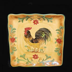 May include: A square ceramic serving dish with a raised rim and a decorative design featuring a rooster with a yellow, black, and green plumage. The rooster is surrounded by green leaves and red and yellow flowers. The dish has a light brown background.