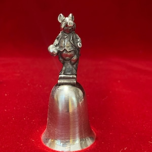 May include: Silver bell with a pig figurine on top. The pig is wearing a suit and holding a small object in its hand.