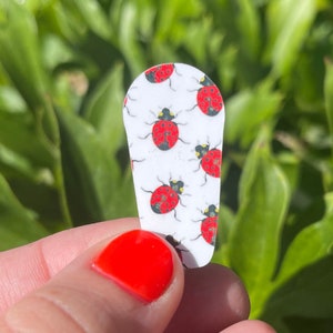 Puede incluir: Una pinza para el pelo blanca con un patrón de mariquitas rojas. La pinza tiene forma de lágrima.