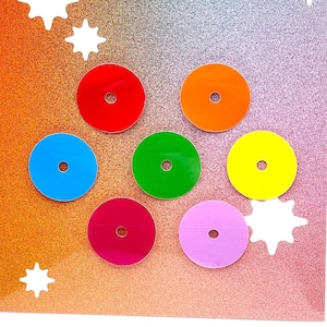 Könnte beinhalten: Ein Satz von sieben bunten runden Aufklebern mit einem Loch in der Mitte. Die Aufkleber sind rot, orange, gelb, grün, blau, rosa und lila. Die Aufkleber sind in einem Kreis auf einem glitzernden Hintergrund angeordnet. Der Text "Bugzy Designs" ist oben im Bild gedruckt.