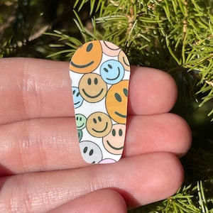 May include: A white oval-shaped sticker with a pattern of colorful smiley faces. The smiley faces are in various shades of yellow, orange, green, and blue.