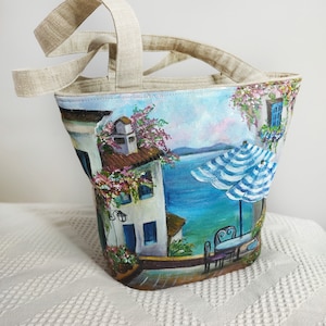Puede incluir: Una bolsa de lona con una escena pintada a mano de un pueblo mediterráneo con un océano azul, edificios blancos y una sombrilla a rayas azules y blancas. La bolsa tiene asas de lino.
