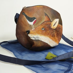 May include: A round brown leather bag with a painted image of a fox and a bird. The bag has a blue strap and a blue mesh drawstring bag.