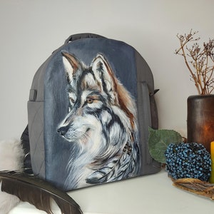 May include: A gray backpack with a painted image of a wolf's head on the front. The wolf is looking to the side and has a gray and white coat with brown accents. The backpack has a zippered pocket on the front and a strap on each side.